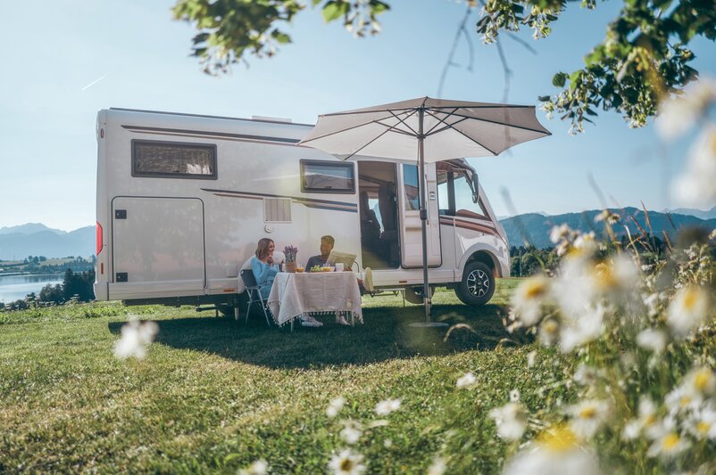 A man and a woman sitting in front of their campervan under a sunshade, drinking coffee.