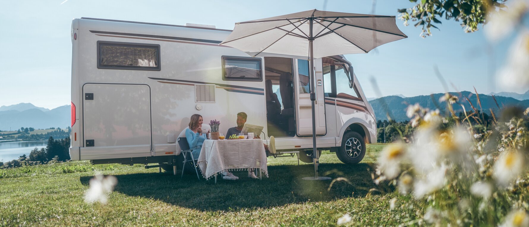 A man and a woman sitting in front of their campervan under a sunshade, drinking coffee.