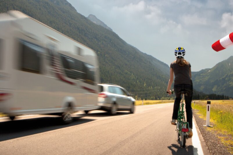 A cyclist rides along the edge of a road while being overtaken by a car towing a caravan. | © AL-KO