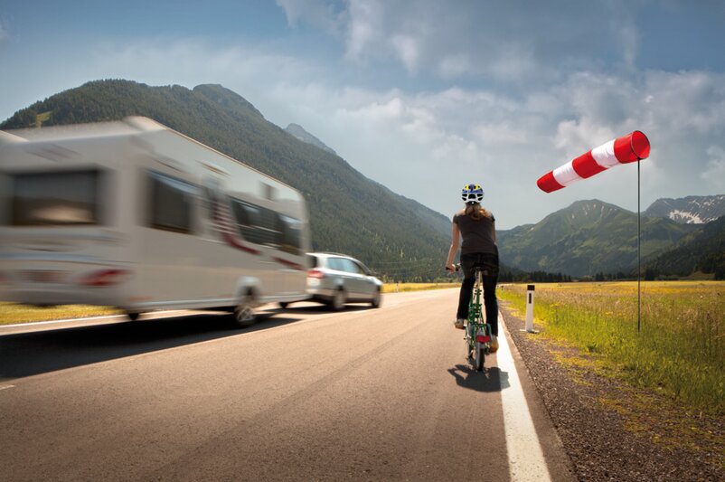A cyclist rides along the edge of a road while being overtaken by a car towing a caravan. | © AL-KO