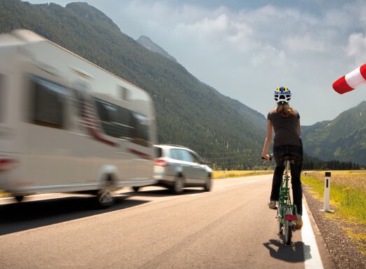 A cyclist rides along the edge of a road while being overtaken by a car towing a caravan. | © AL-KO