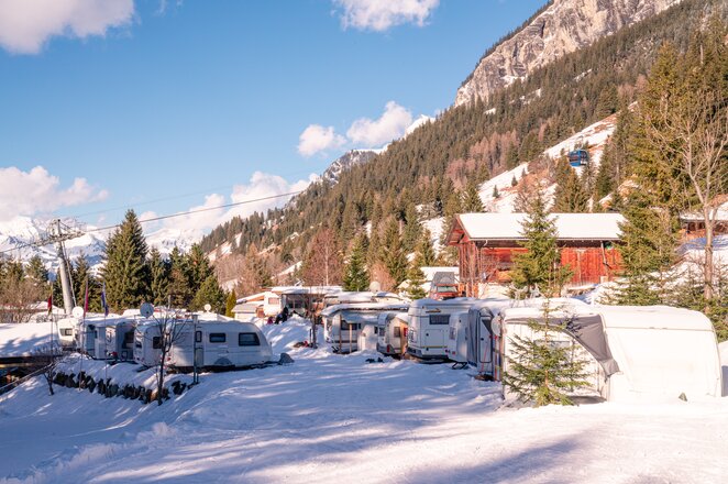 A snow-covered mountain campsite with several caravans; a ski lift is visible in the background.