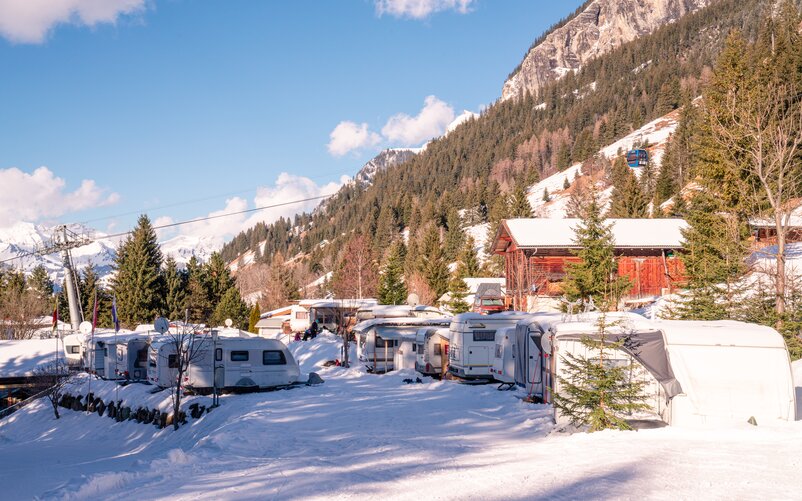A snow-covered mountain campsite with several caravans; a ski lift is visible in the background.