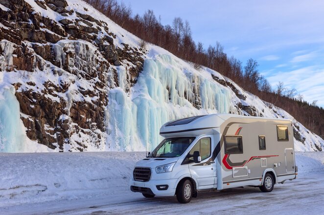 A camper van parked on a snow-covered lot in front of a rock wall with icicles.