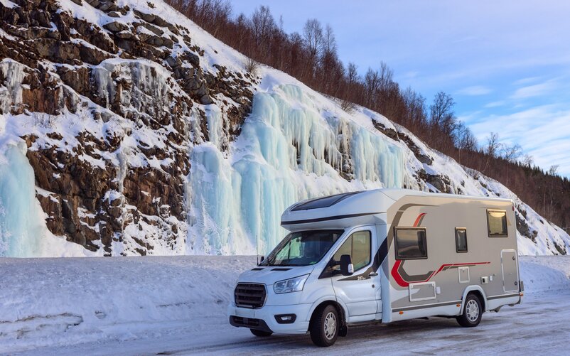 A camper van parked on a snow-covered lot in front of a rock wall with icicles.