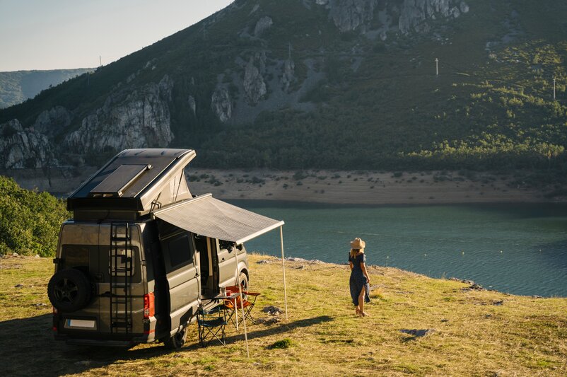 Ein Frau steht in einem Kleid und mit einem Hut neben einem Campingfahrzeug an einem Fluss, der vor einem Berg vorbeifließt.