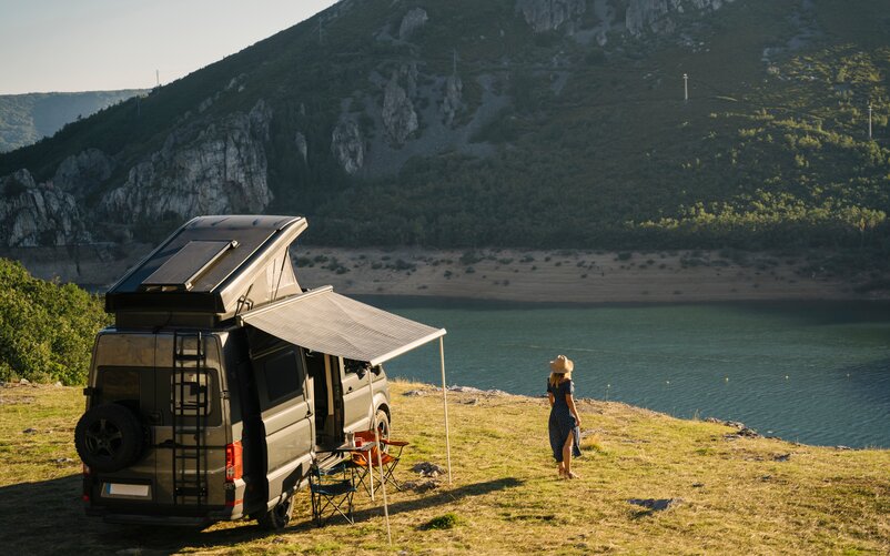 Ein Frau steht in einem Kleid und mit einem Hut neben einem Campingfahrzeug an einem Fluss, der vor einem Berg vorbeifließt.