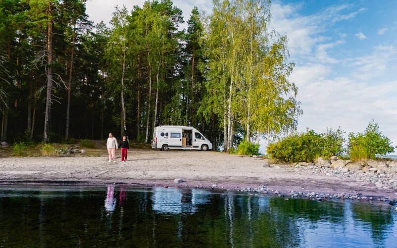 Ein Paar steht am Ufer, im Hintergrund sind ein weißes Wohnmobil und Bäume zu sehen.