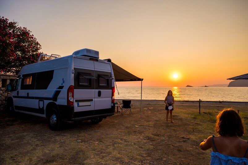 Two children play frisbee on the beach at sunset beside a motorhome.