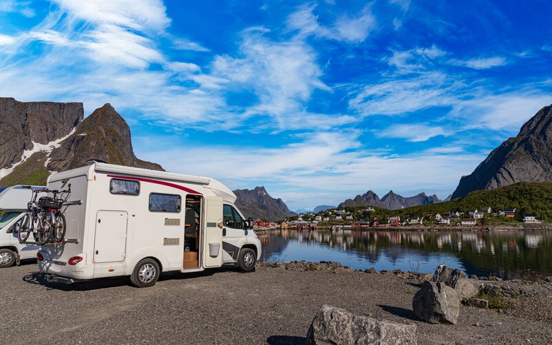 A motorhome is parked by the shore of a fjord. Scandinavian houses can be seen on the opposite shore.