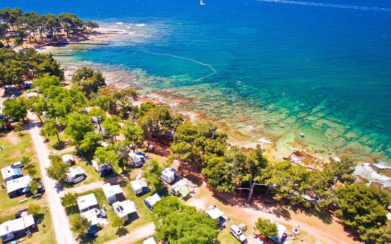 A campsite with several motorhomes among trees by the sea, photographed from above.